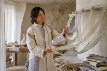 Landscape environmental portrait of an anonymous gesture archivist guiding translucent fabric ribbons inside a bright ivory and sage textile atelier.