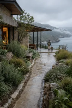 Anonymous traveler with plain luggage pausing on a rain-washed limestone terrace overlooking a misty coastal inlet and layered cliffs.