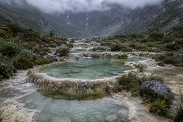 Rainy highland basin with shallow jade-tinted wetland pools cascading across pale natural stone terraces beneath misty mountain walls.