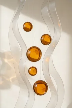 Abstract studio composition of amber glass spheres suspended among translucent folded acrylic ribbons on a warm off-white background.