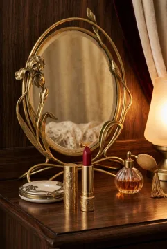 Crimson lipstick beside a pearl powder compact and glass perfume atomizer on an Art Nouveau vanity with a floral brass mirror and warm retro lighting.