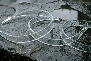 Translucent glass ribbons curve across dark wet stone with rain droplets, soft natural light, and subtle reflections in a photorealistic abstract composition.