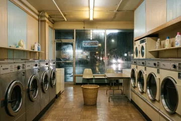 Rainy 1980s coin laundry at midnight with chrome washing machines, empty plastic chairs, a laundry basket, and reflections across wet storefront windows.