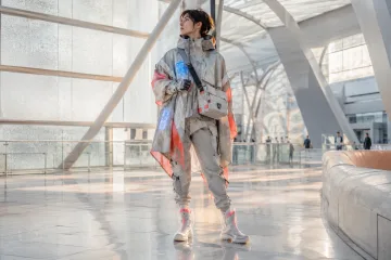 Anonymous near-future urban courier in an adaptive geometric smart-textile coat standing inside a bright metropolitan transit atrium.